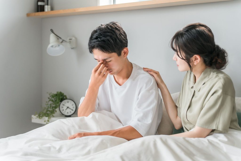 ベッドに座って目を押さえている男性と寄り添う女性